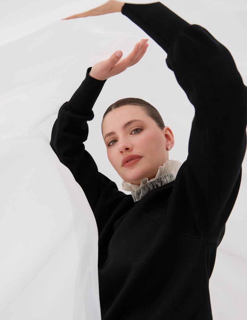 Woman in black sweater with frilly white collar, poses in front of a bright white background, holding white fabric overhead.