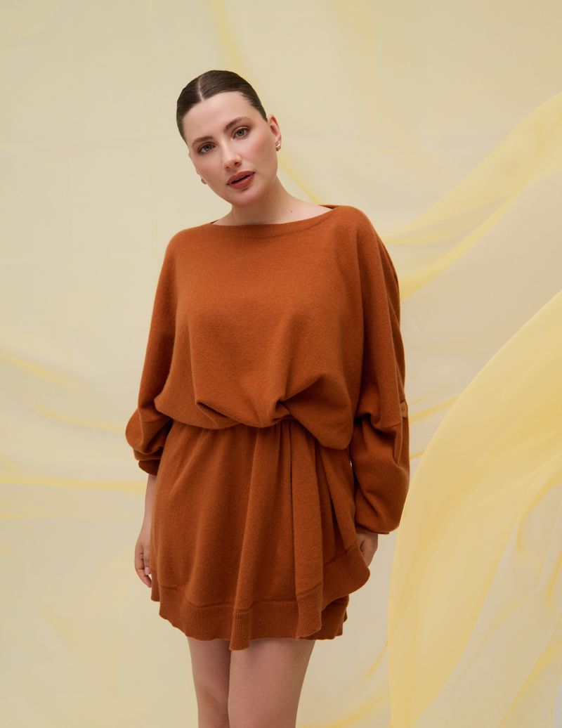 Elegant brunette woman posing in a warm terracotta sweater dress against a soft yellow background with sheer fabric overlay.
