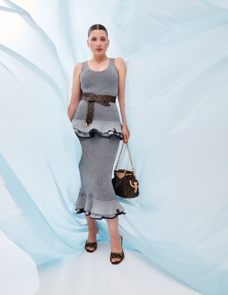 Elegant adult female poses in a studio against a flowing blue backdrop wearing a matching gray dress, brown belt, and carrying a bucket bag.