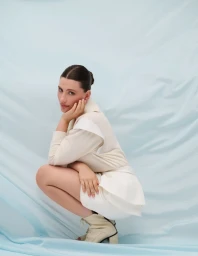 Model crouching wearing a cream outfit and matching ankle boots against a pale blue fabric backdrop.