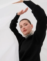 Model posing in a high-neck black sweater with ruffled collar holding sheer white fabric against a white background.