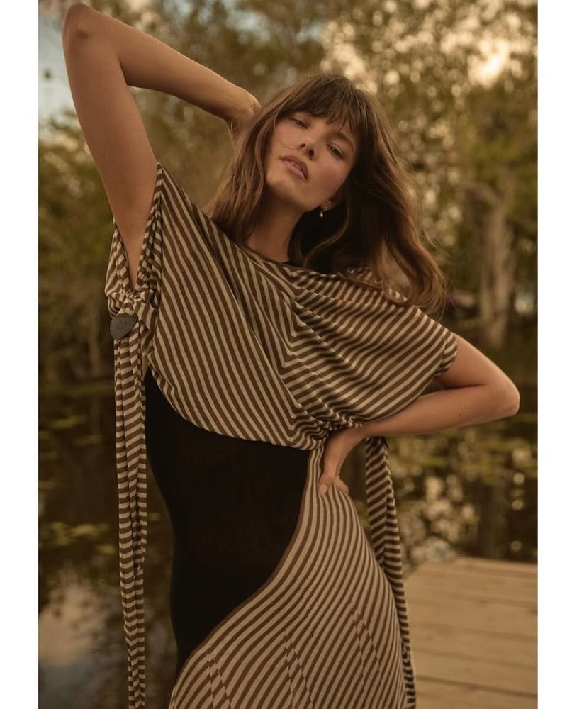 Woman in striped dress poses outdoors with hand on hip in nature background, fashion, beauty, and style, looking at the camera.