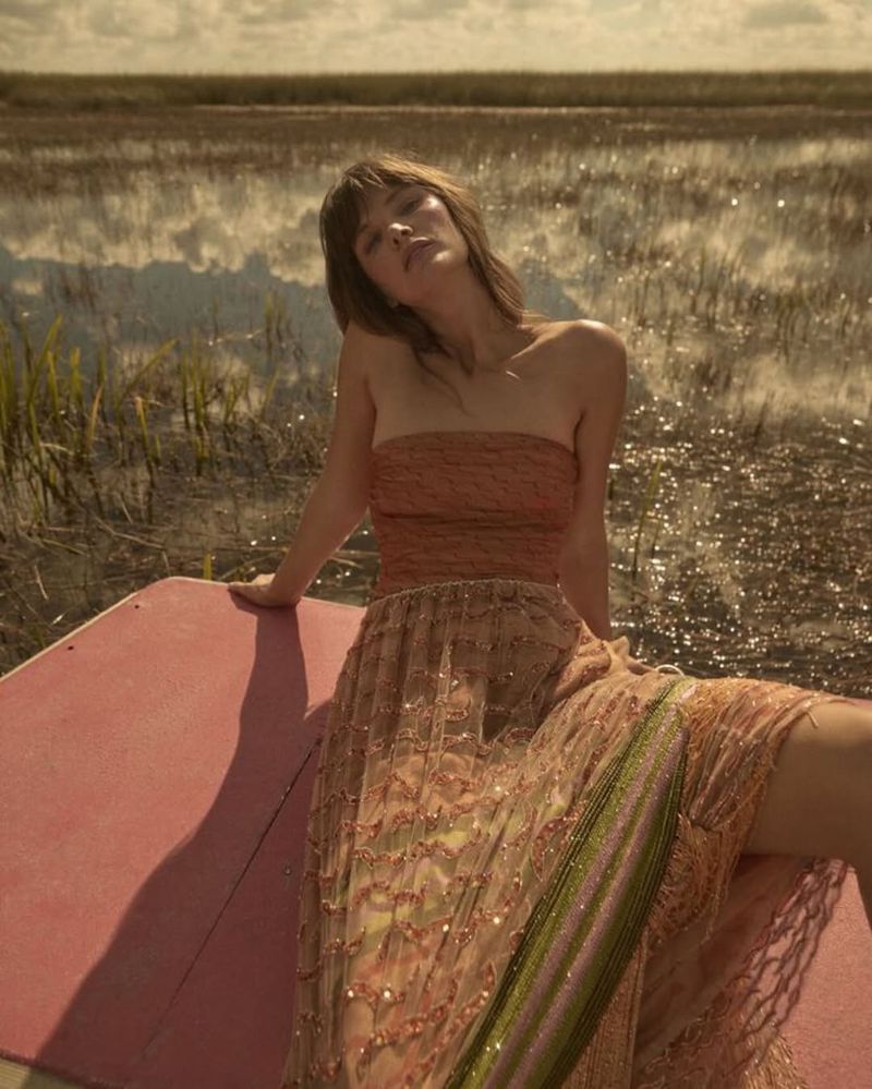 A woman in a strapless top and long patterned skirt is seated on a pink surface with a marsh and sky in the background.