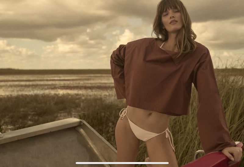 Woman poses in a brown crop top and bikini bottoms on a boat with a field and cloudy sky in the background, fashion shot, outdoor setting.