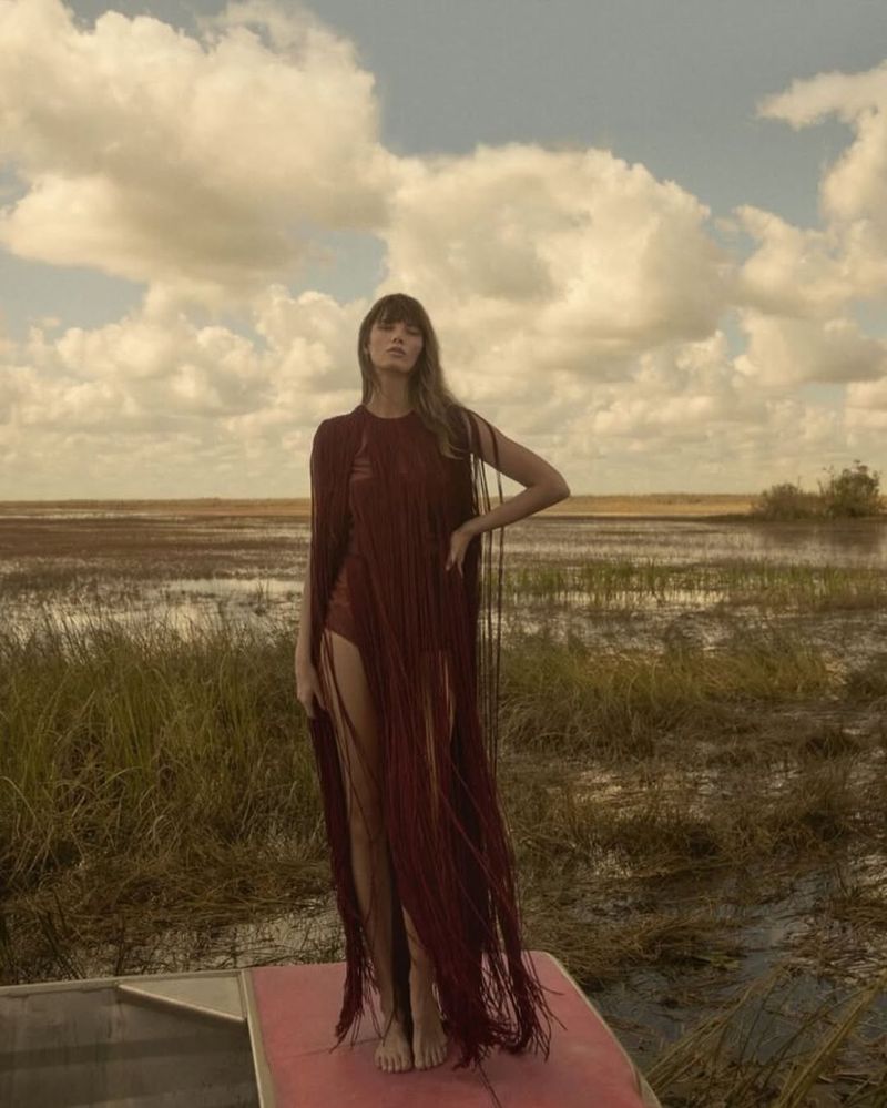 A woman in a fringed garment stands on a boat in a marshy landscape under a cloudy sky, posing for a fashion editorial.
