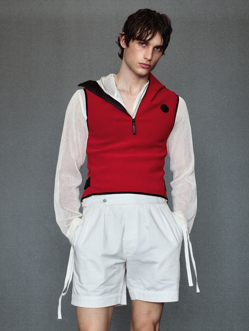 Male model poses in a red sleeveless hooded top and white shorts against a gray backdrop in a studio setting for a fashion editorial.