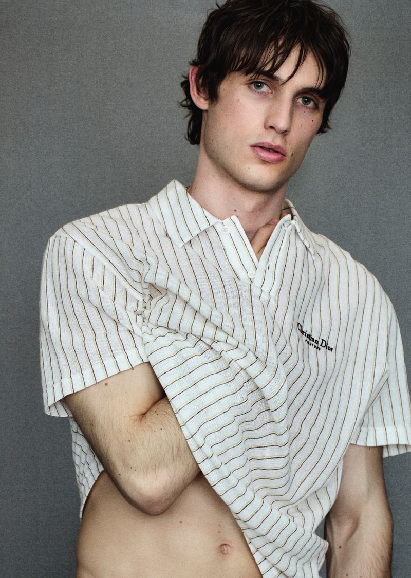 Young man with dark hair in a white pinstripe short-sleeve polo shirt lifting the hem against a grey studio backdrop.