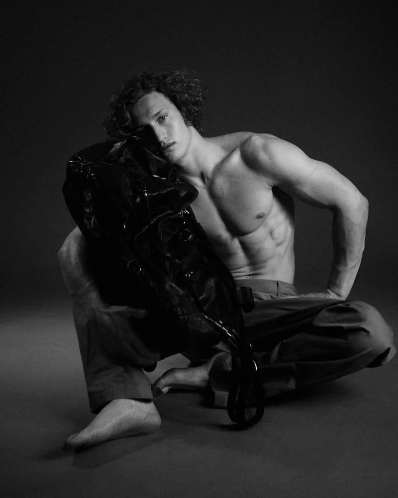 Athletic man with curly hair poses shirtless while crouching with a shiny black coat in a studio with dark gray backdrop.