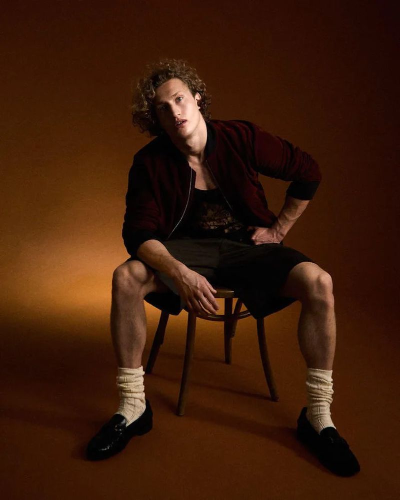 Stylish young man sits on wooden chair in studio, wearing a bomber jacket, shorts, socks and loafers against a brown backdrop.