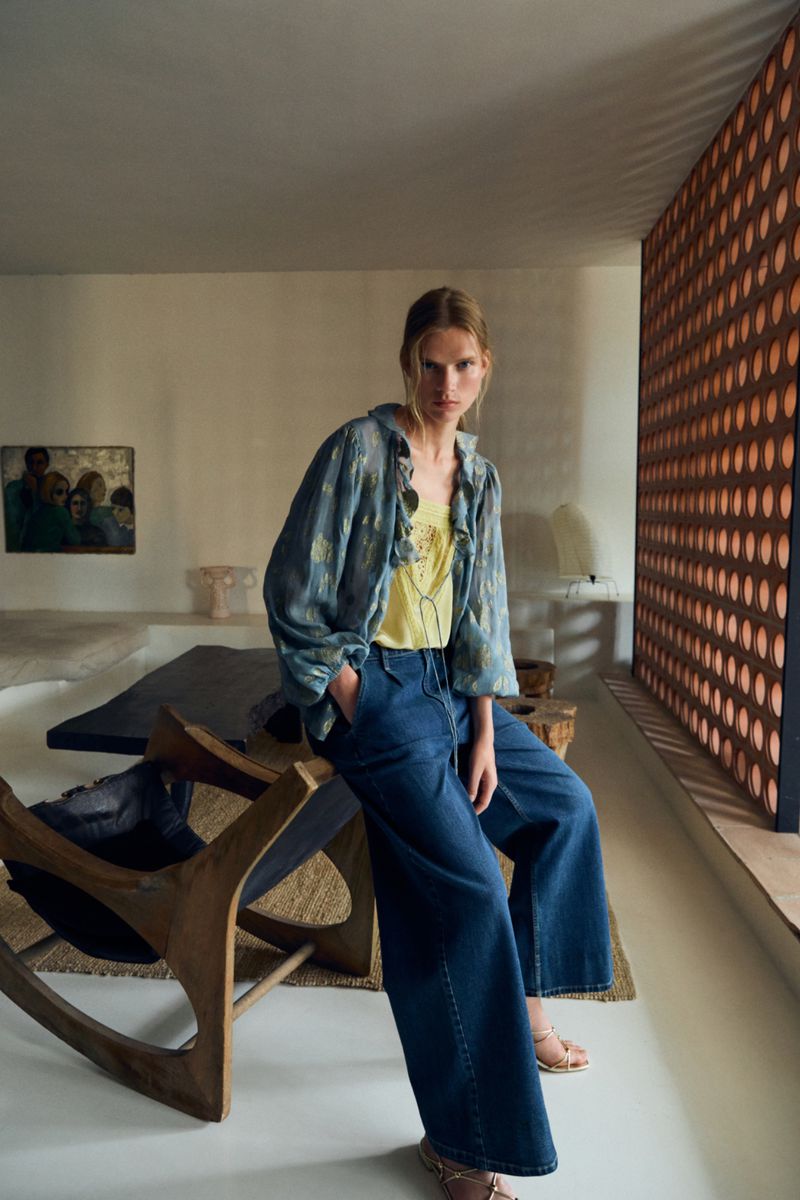 A woman in a blue floral jacket and dark denim wide-leg jeans poses in an indoor setting with neutral tones and warm lighting.
