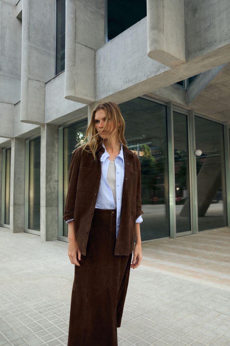 Adult woman in a corduroy jacket and maxi skirt posing in an urban environment with modern concrete architecture and large windows.