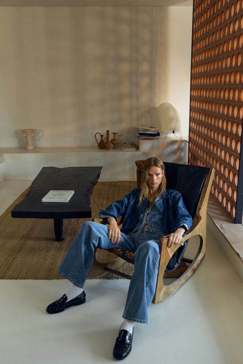 Adult woman poses indoors wearing denim jumpsuit, jacket, loafers, white socks, and sits in a wooden chair in a stylish interior.