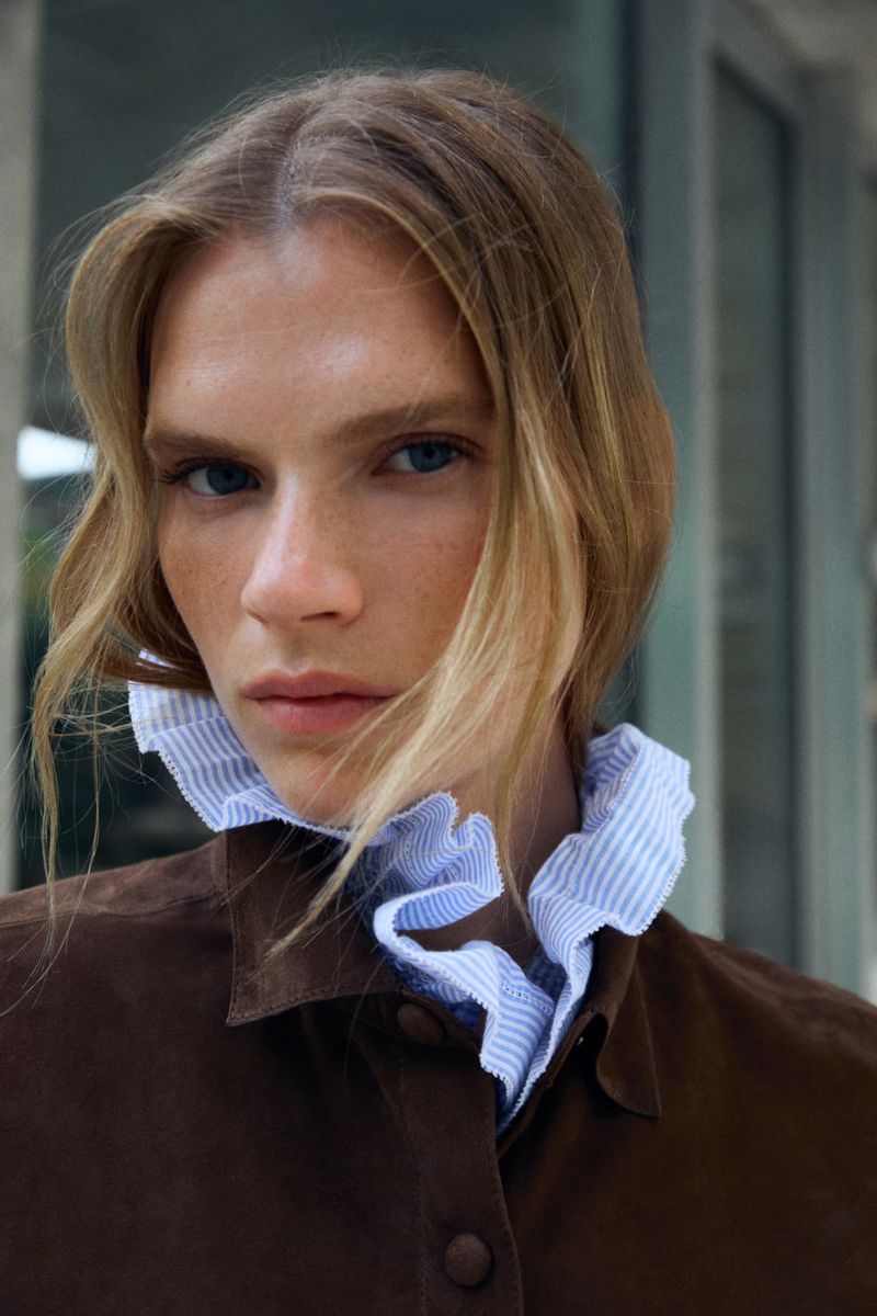 Close-up portrait of a fair-skinned adult female with blonde hair wearing a brown jacket and a frilly-collared blue-striped shirt.