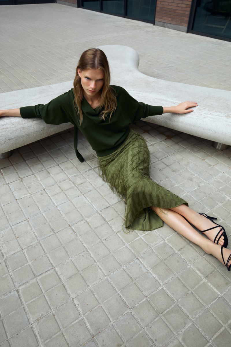 A woman poses outdoors in a green sweater and textured skirt, seated on a concrete bench with arms extended, looking directly at the camera.