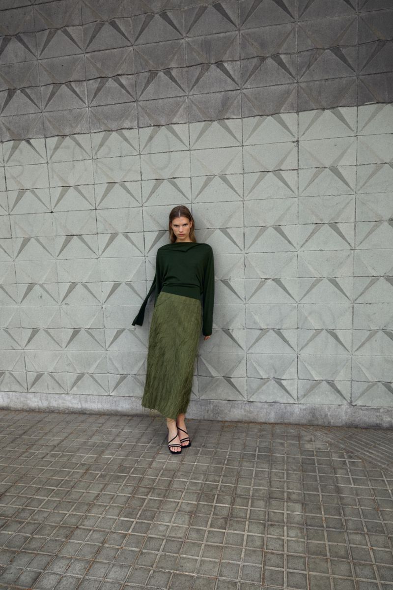 A slender woman stands in front of a textured concrete wall, wearing a long-sleeved dark green top and a textured, knee-length olive green skirt.