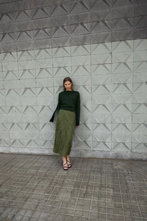 A slender woman stands in front of a textured concrete wall, wearing a long-sleeved dark green top and a textured, knee-length olive green skirt.