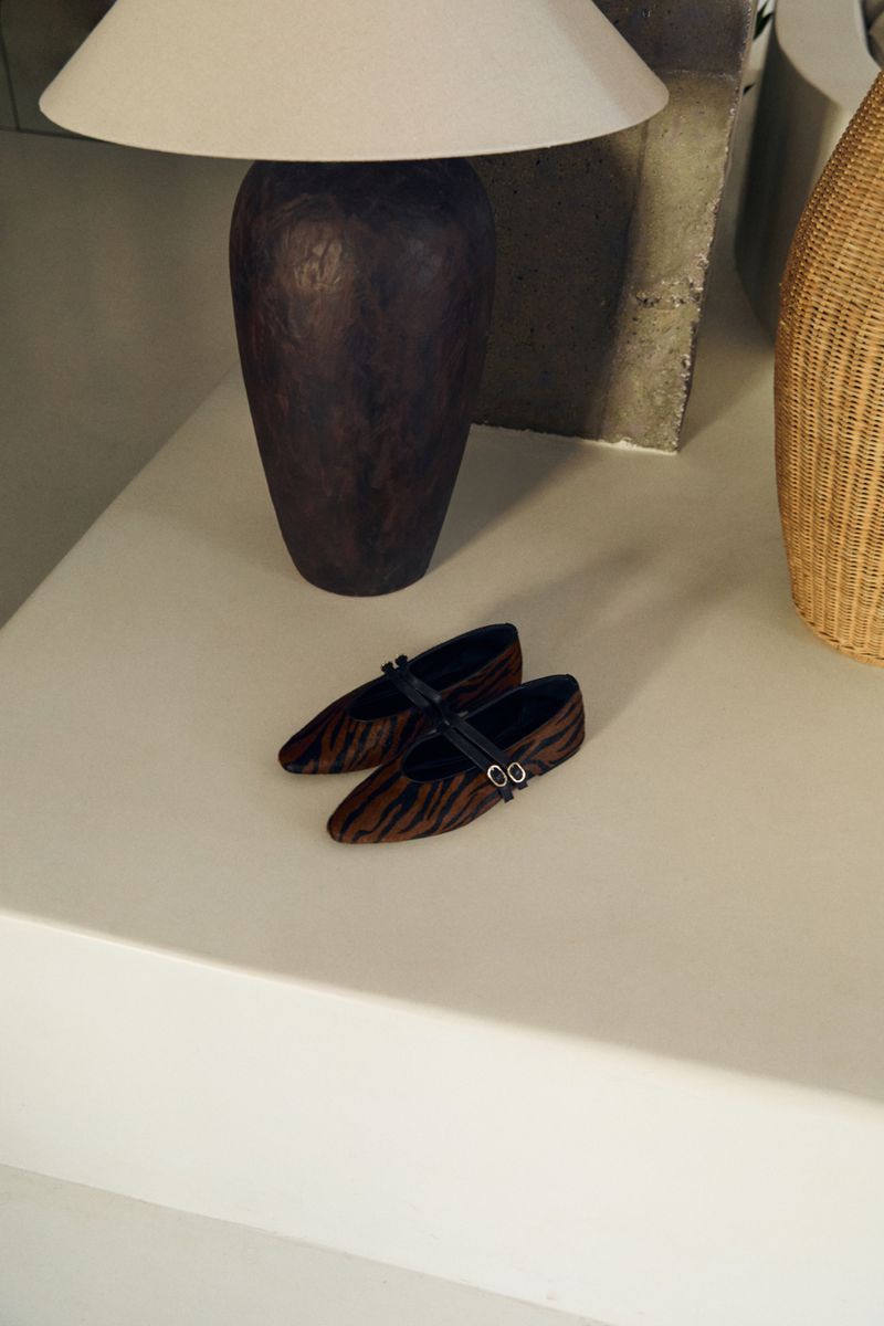 Pair of tiger-striped Mary Jane shoes with black straps displayed on a white surface, with a lamp and basket in the background.