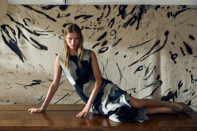 Fashion shot of a blond adult woman posing on a dark wooden surface. She is wearing a dress with an abstract design on it. 
