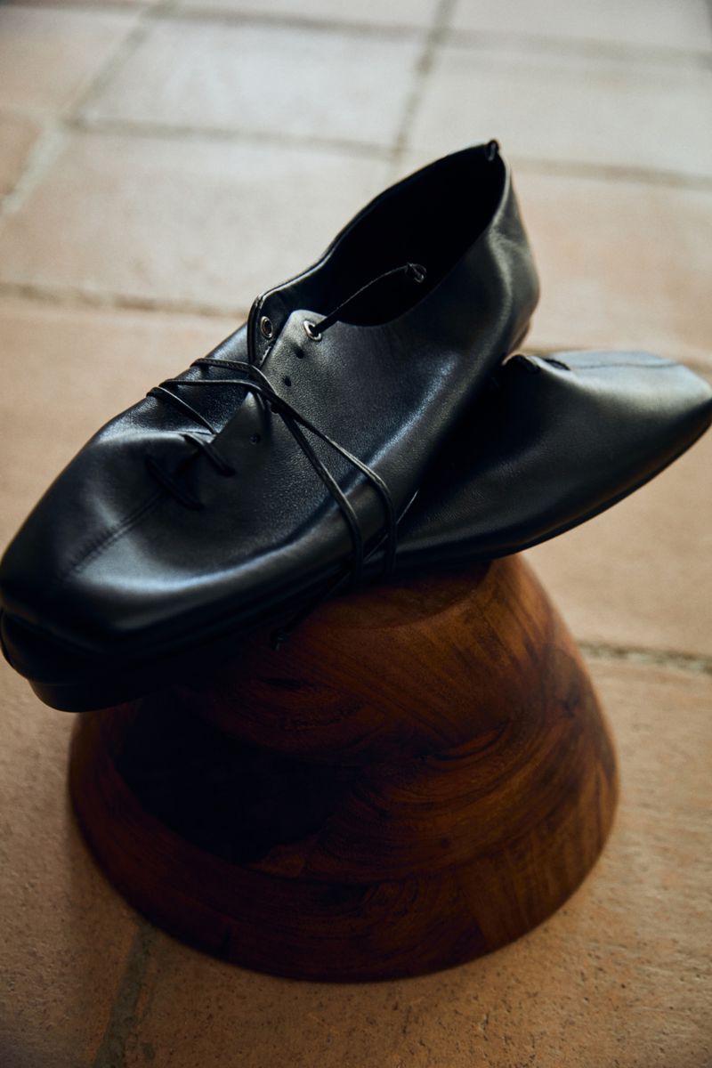 Elegant black leather shoes displayed on a dark wood pedestal against a textured stone tile backdrop. Fashion footwear product photography.
