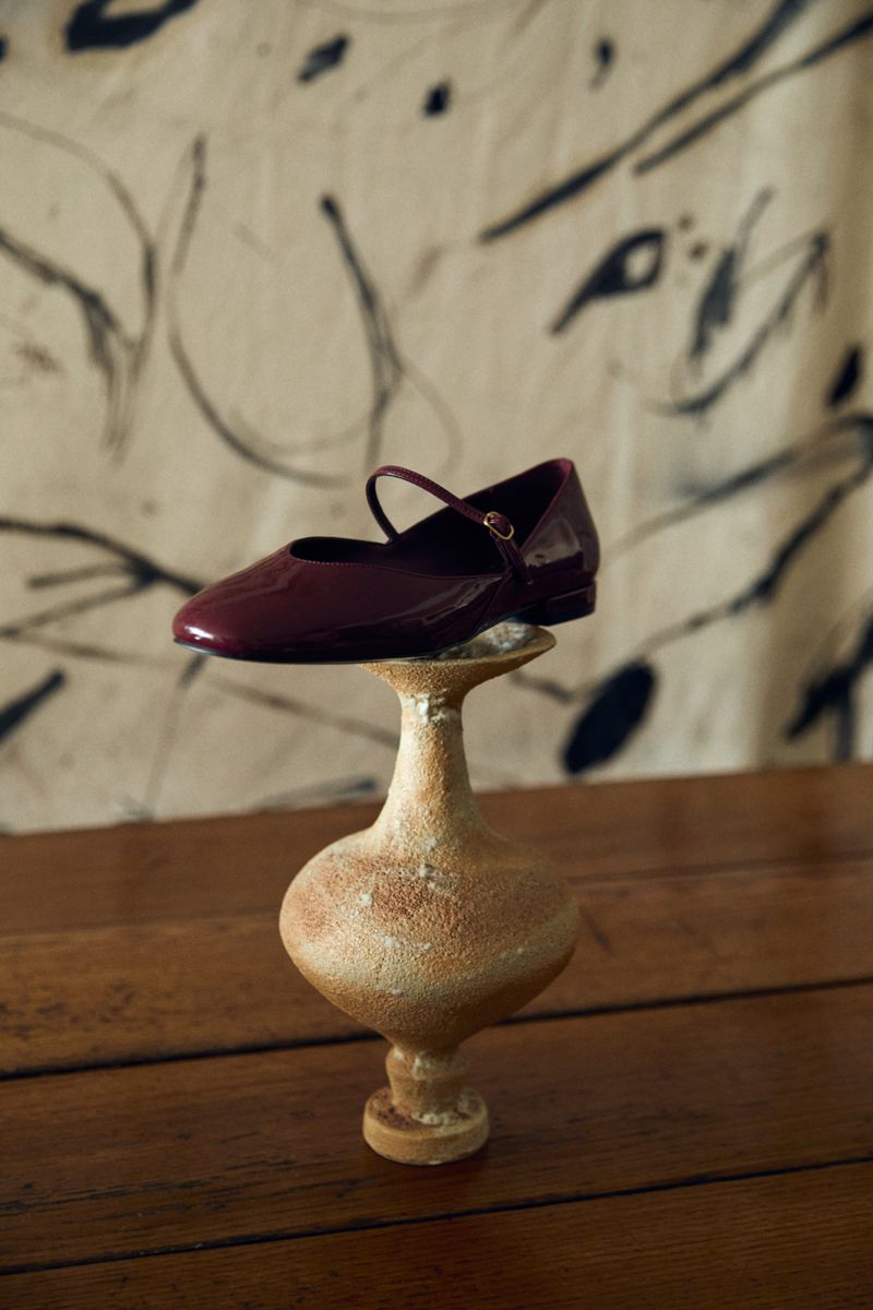 Burgundy Mary Jane shoe rests atop a textured tan pedestal against an abstract art backdrop in a fashion-forward still life composition.