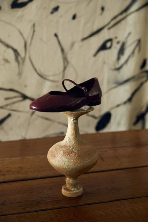 Burgundy Mary Jane shoe rests atop a textured tan pedestal against an abstract art backdrop in a fashion-forward still life composition.