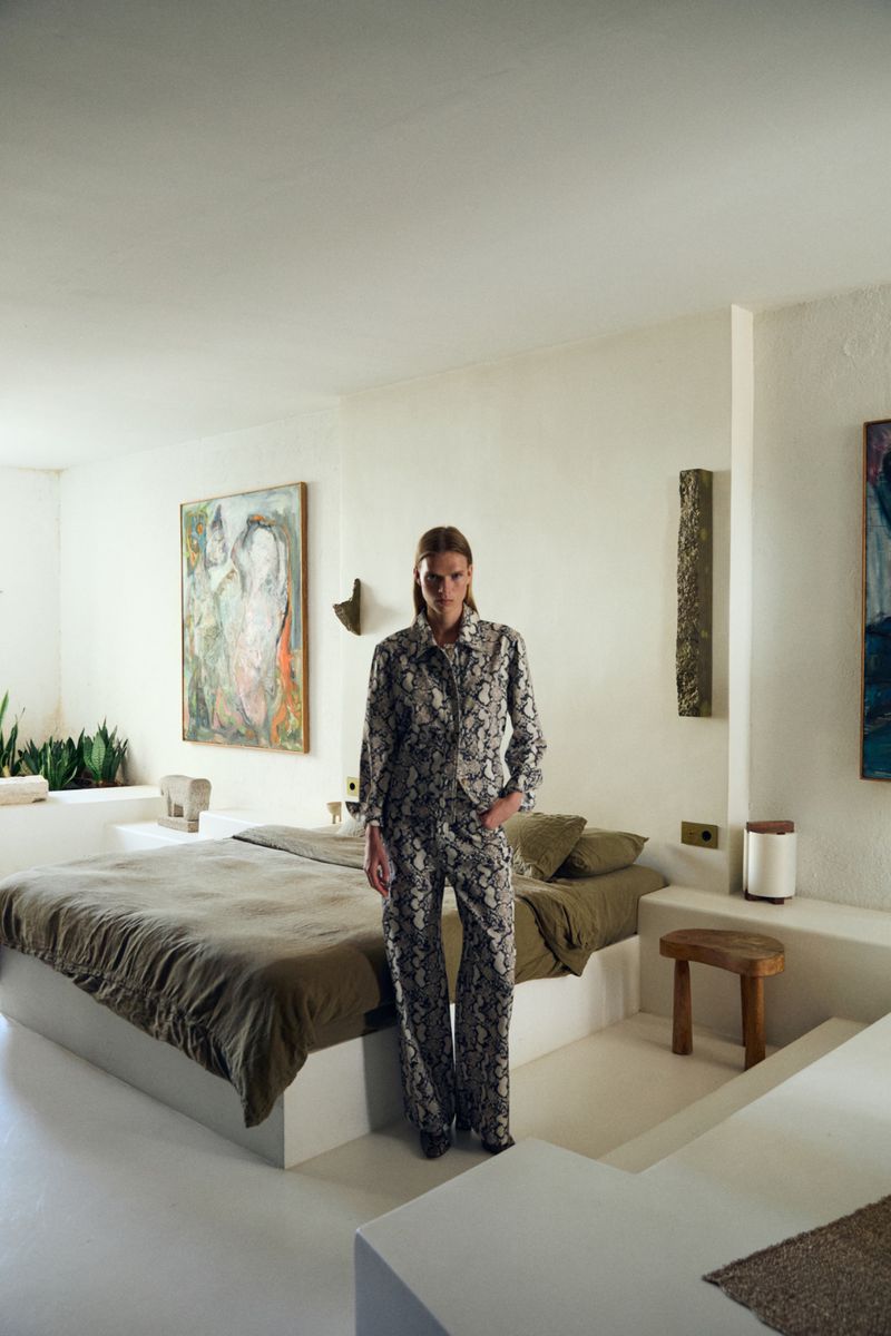 Fashionable woman in patterned pajamas stands in a minimalist interior bedroom with white walls, art, olive green bed, and a stool.
