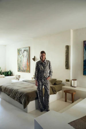 Fashionable woman in patterned pajamas stands in a minimalist interior bedroom with white walls, art, olive green bed, and a stool.