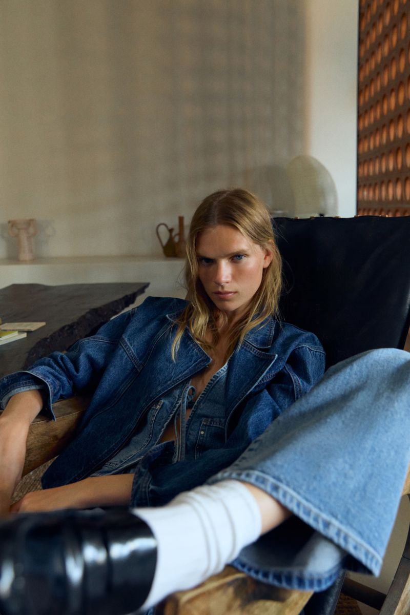 Blonde model in denim jacket and jeans, seated in modern chair with white socks and black loafers, studio lighting, interior background.