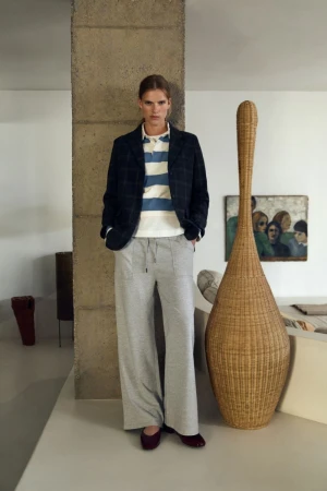 Standing adult wearing a dark blazer, striped polo shirt, and wide-leg sweatpants, posed in an interior setting with neutral colors.