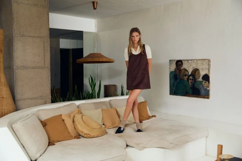 Model Sara Eirud standing on a large cream sofa wearing a brown pinafore dress over a white collared shirt.