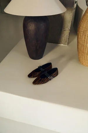 Tiger print Mary Jane flat shoes resting on a cream architectural ledge beside a dark ceramic table lamp.