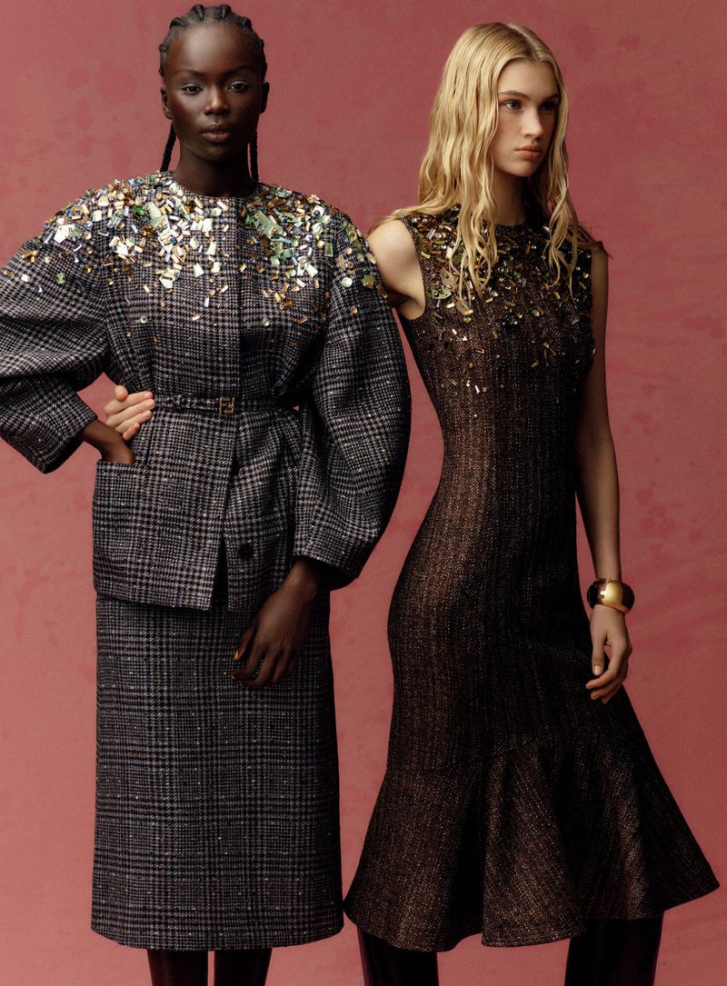 Two fashion models pose against a warm rose backdrop wearing embellished designer outfits including a grey plaid skirt suit and dark midi dress.