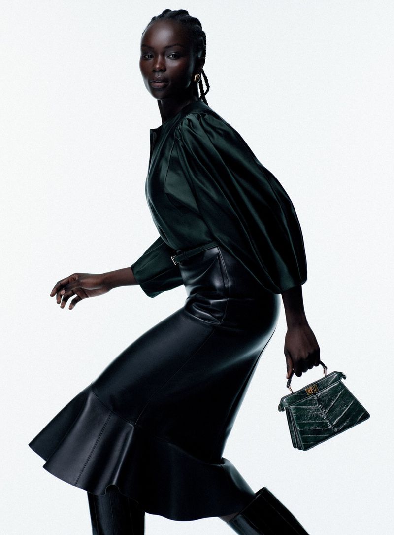 A Black female fashion model in a green blouse and black skirt holds a green handbag against a bright white background.