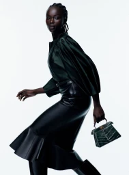 Black woman in a dark green satin blouse and black flared leather skirt walking with a green handbag against a white background.