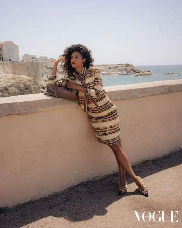 Model Melodie Vaxelaire in a patterned tweed skirt suit leaning against a coastal wall with a brown handbag.