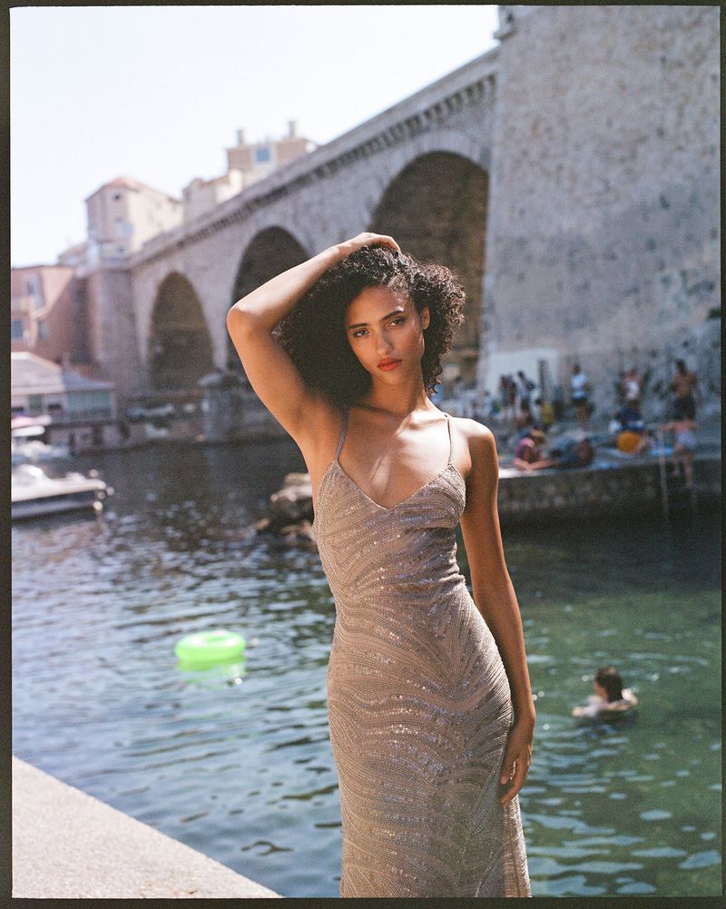Woman in a shimmering dress poses in front of a stone bridge and a body of water on a sunny day. Outdoor fashion photography.