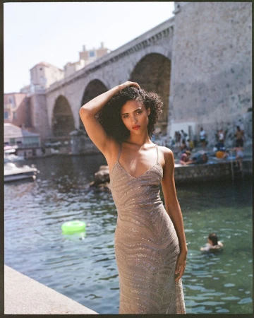 Woman in a shimmering dress poses in front of a stone bridge and a body of water on a sunny day. Outdoor fashion photography.