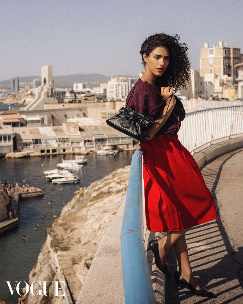Fashion shot of a woman in a red skirt and maroon top, posing on a bridge overlooking a coastal city, with the Vogue logo visible.