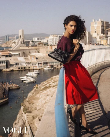 Fashion shot of a woman in a red skirt and maroon top, posing on a bridge overlooking a coastal city, with the Vogue logo visible.