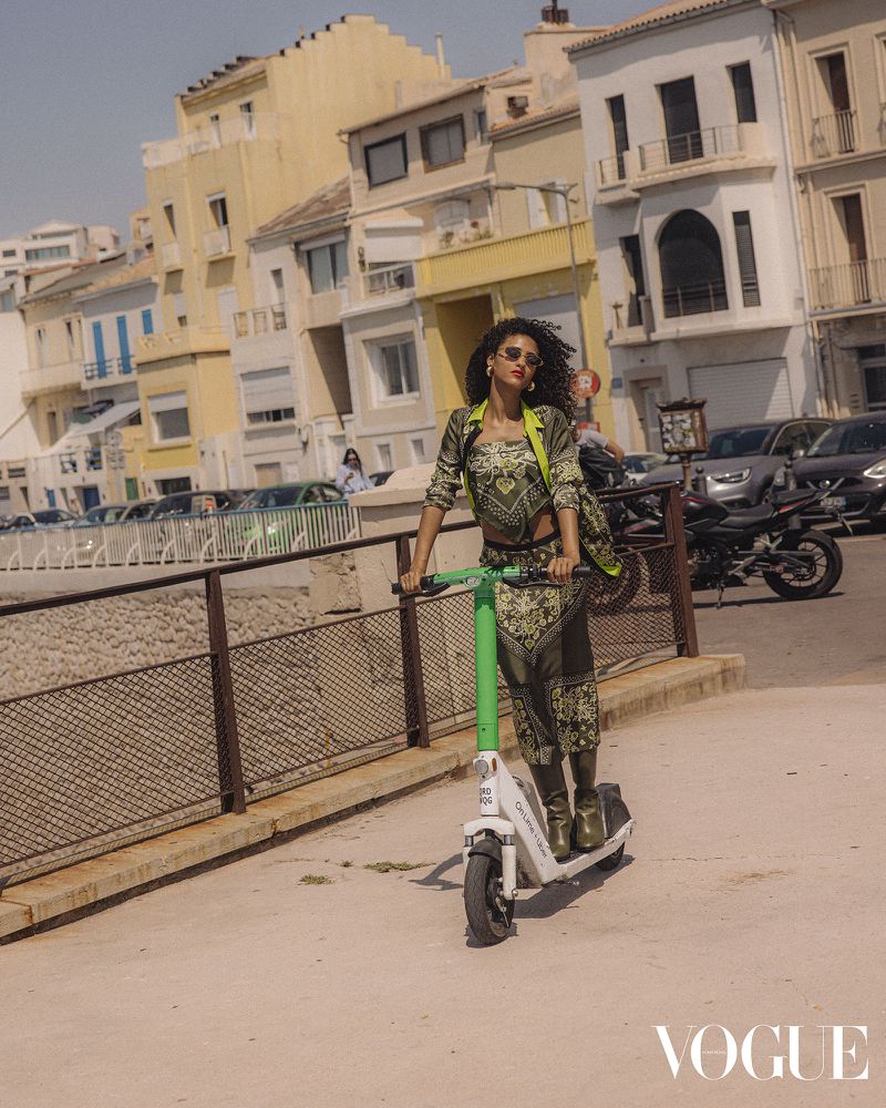 Woman in patterned suit riding an electric scooter on a sunny city street, with buildings and parked vehicles in the background.