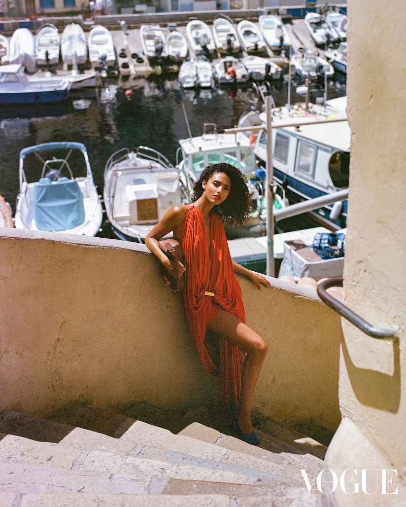 Fashion shot of a woman in a red dress on steps near boats, featuring a tan wall, and blue water with boats in a harbor.