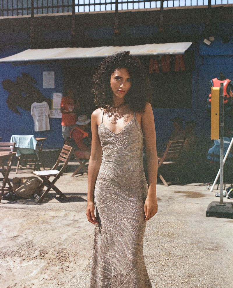 Adult female model with curly hair wears an elegant evening gown outdoors with a building and a lifeguard rescue station in the background.
