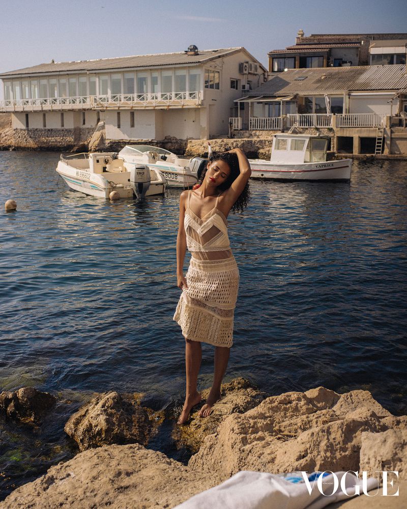 Adult woman in a crocheted dress poses barefoot on rocks by the sea, with boats and buildings visible in the background under sunny skies.
