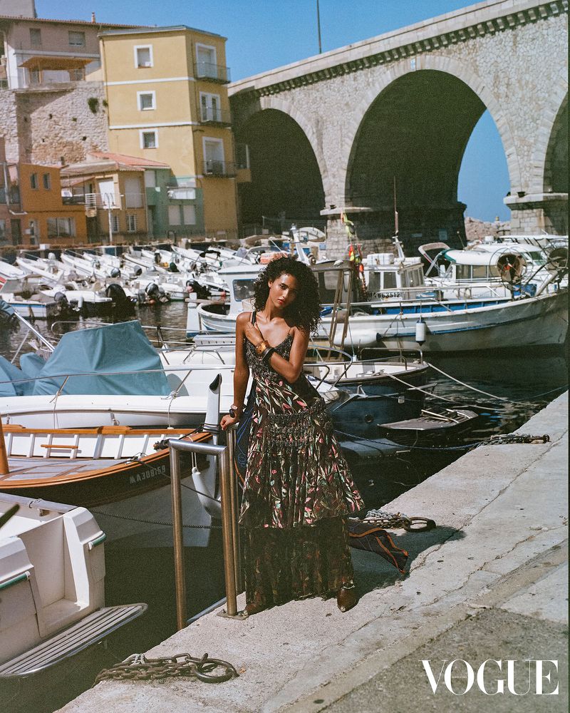 A woman in a long dress poses near boats on a sunny day with stone bridge and buildings in the background in a fashion editorial.