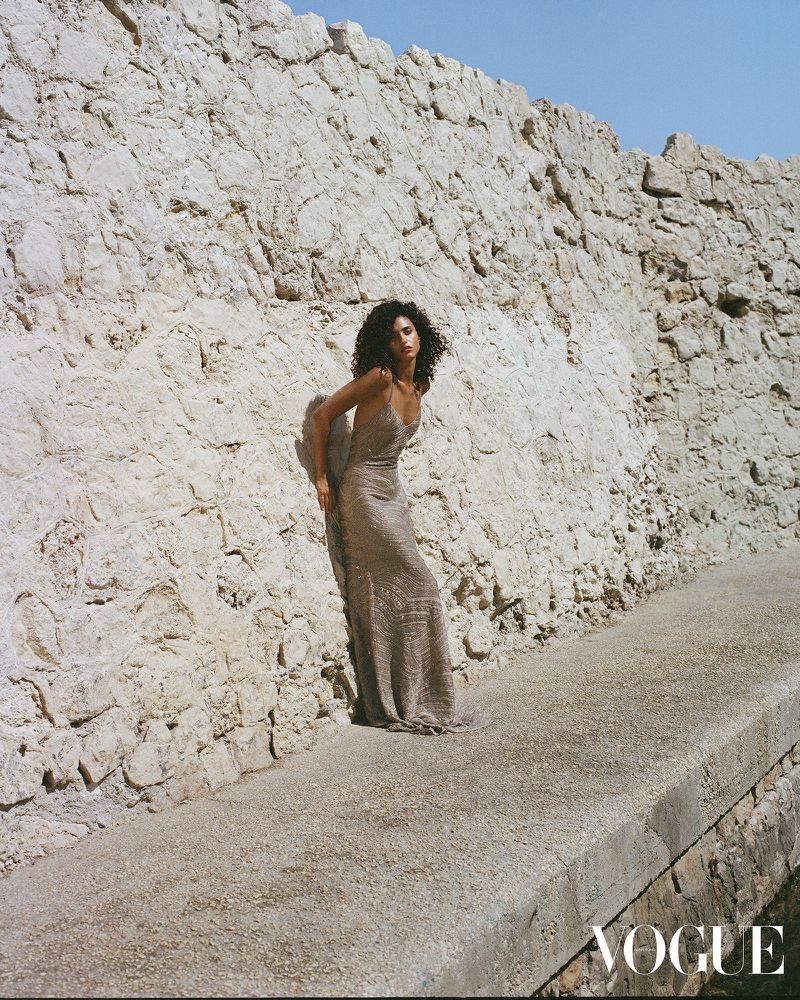 An adult female model with curly hair poses outdoors in a dress against a stone wall on a sunny day for a fashion editorial.