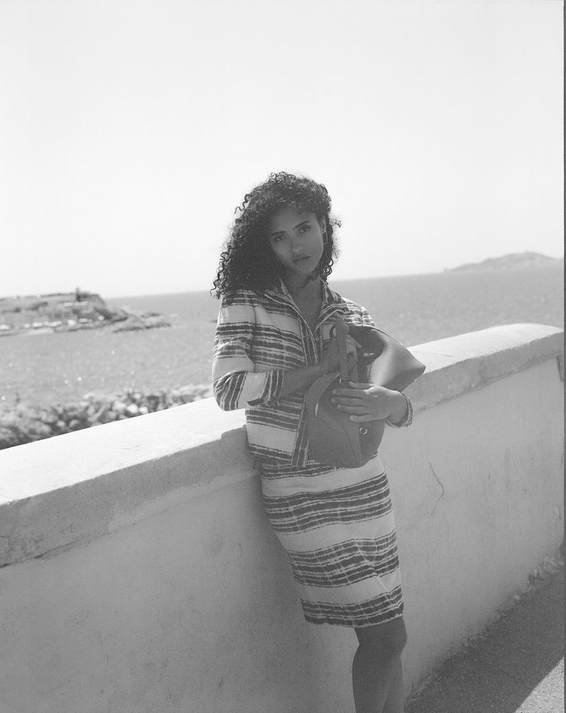 A curly-haired woman in a striped jacket and skirt, posing outdoors with a handbag, against a seaside backdrop on a sunny day.