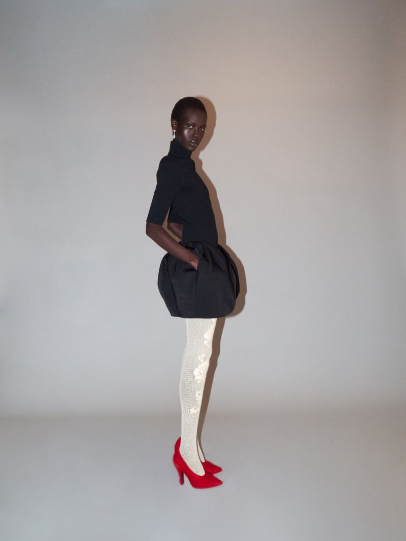 Woman poses in a studio wearing a black dress with a puffy skirt, white patterned tights, and red high heels against a neutral backdrop.