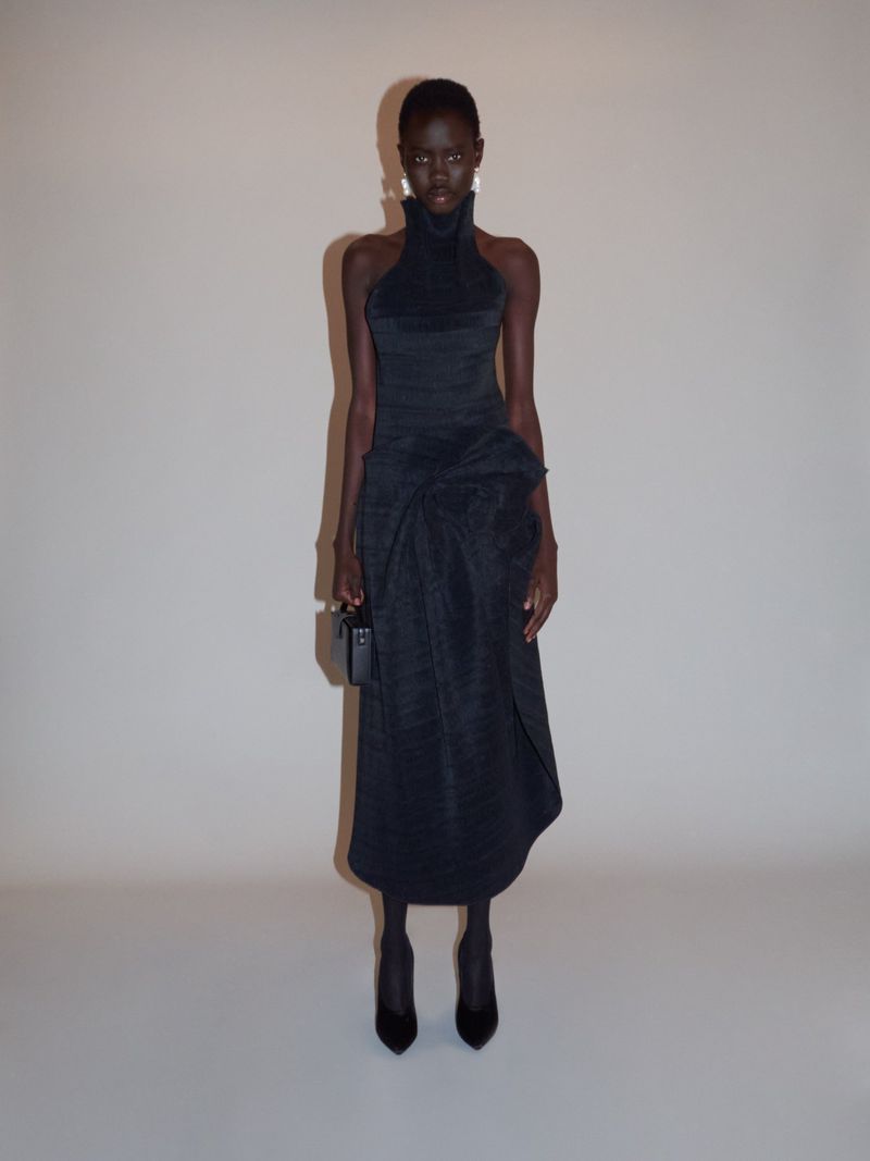 Full-length shot of a Black woman in a black high-neck dress, holding a small black box purse, standing in a neutral studio setting.