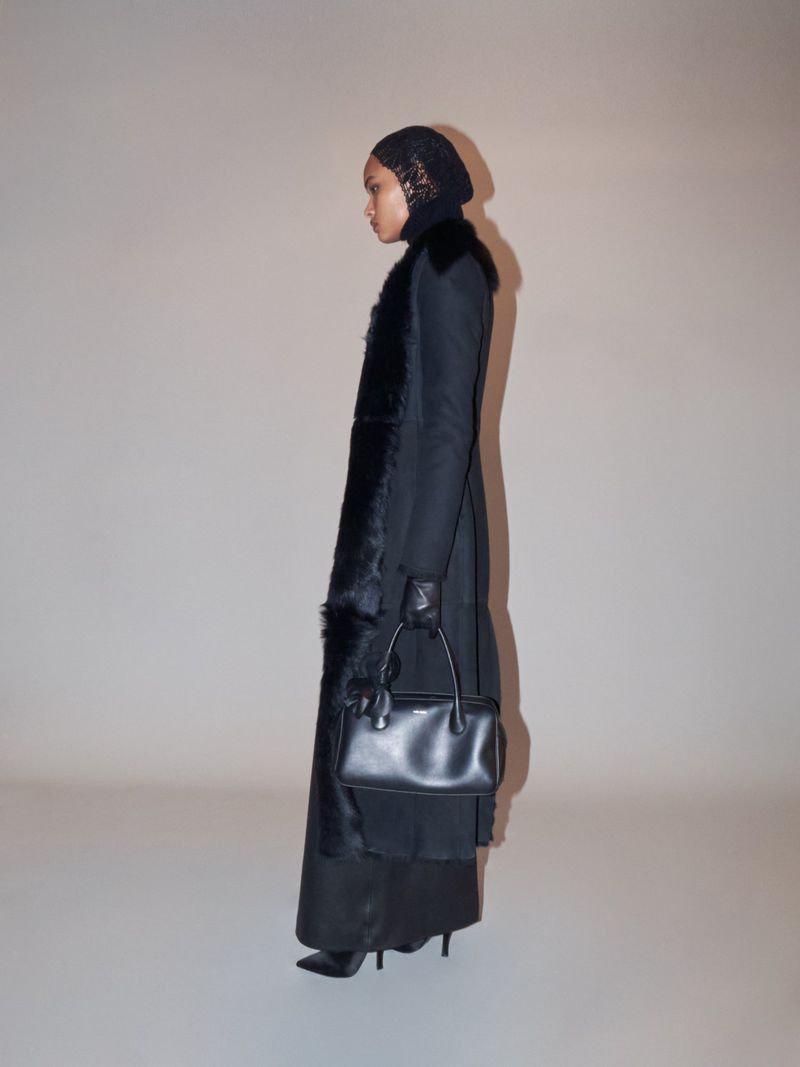 A slender adult female poses indoors in a long black coat with fur trim, a black headdress, black gloves, and carrying a small black handbag.