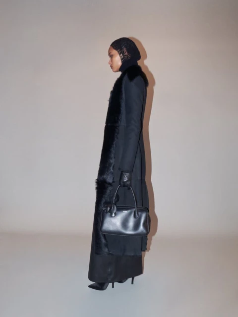A slender adult female poses indoors in a long black coat with fur trim, a black headdress, black gloves, and carrying a small black handbag.